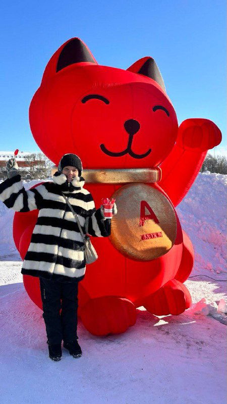 Большая красная фигура кота-талисмана на фестивале, рядом посетитель в полосатом пальто на фоне ясного зимнего неба и снега.