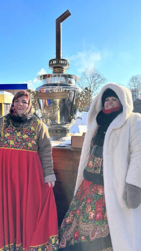 Две женщины в ярких традиционных сарафанах у самовара: уличная сцена фестиваля, чайники, народные костюмы и зимняя атмосфера.