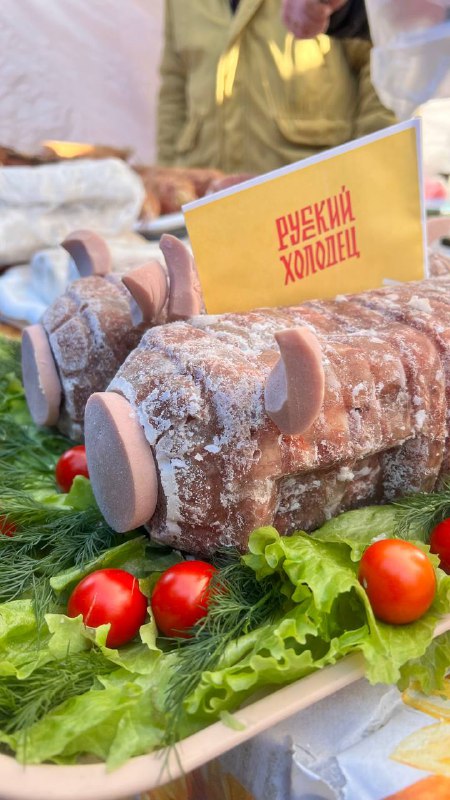 Крупный план холодца, оформленного на подносе в виде бревнышка с зеленью и табличкой «Русский холодец», сервировка и элементы декора.