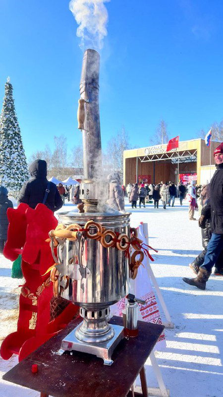 Большой самовар на подставке с клубящимся паром и гирляндами, рядом сувениры и декоративные элементы фестивальной зоны на снегу.