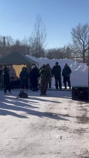 Палатки и очереди у прилавков фестиваля на зимней площадке: посетители в шапках и куртках, заметны тени и яркое солнце.