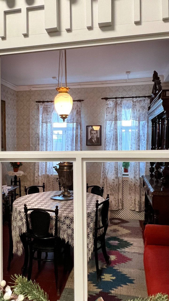 Интерьер комнаты в доме‑музее: стол с ажурной скатертью, самовар и старинная мебель, вид через разделённое окно‑перегородку, домашняя атмосфера экспозиции.
