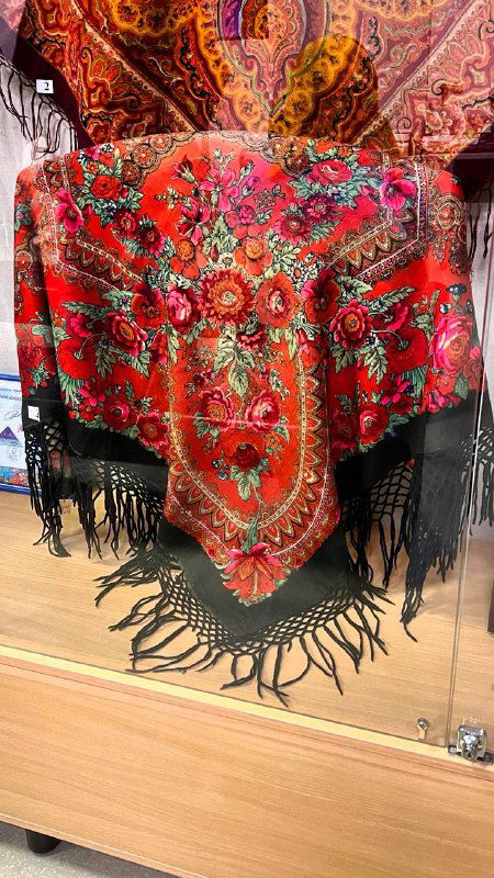 Большой традиционный павловопосадский платок с ярким орнаментом и чёрной бахромой, развешенный в экспозиций под стеклом на деревянной подставке.