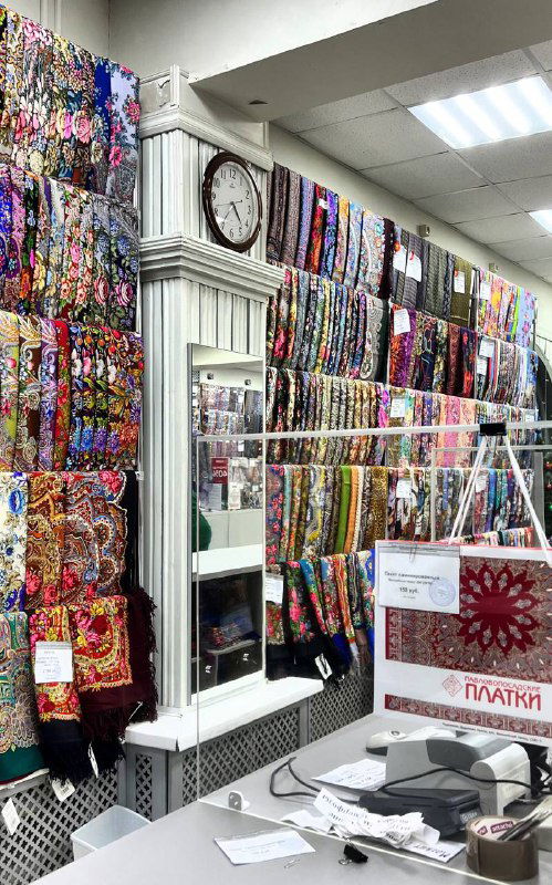 Магазин мануфактуры: полки, заполненные яркими платками и шалями разных цветов и узоров, касса и рекламная табличка на прилавке.