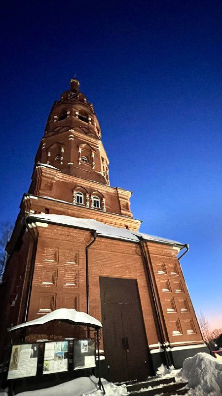 Колокольня и башня храма в тонкой вечерней подсветке: высокое здание с традиционной архитектурой и снежными акцентами у подножия.