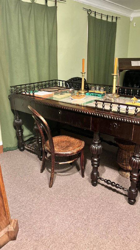 Письменный стол в музее Толстого с подсвечником и предметами быта на зелёном фоне штор — атмосфера рабочего кабинета XIX века.