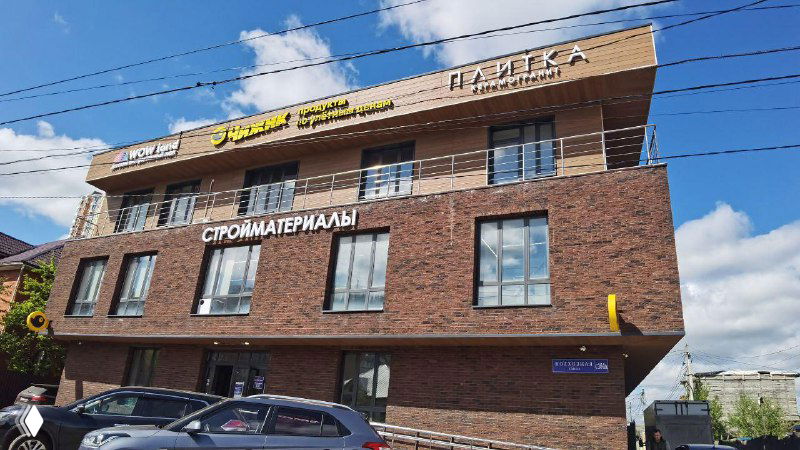 Крупный план фасада того же коммерческого здания с вывесками стройматериалов и витринами магазинов на фоне ясного неба и припаркованных машин.