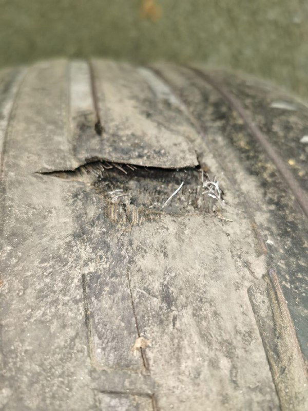 Крупный план сильно повреждённой автомобильной шины с вырванным куском резины и видимым изломом протектора на асфальте.