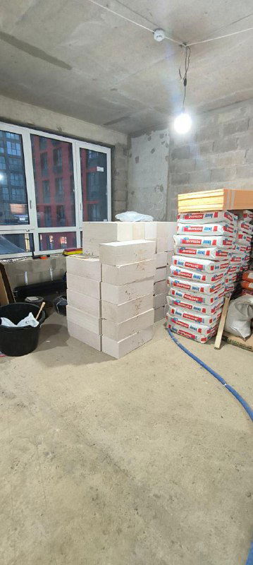 Интерьер будущих студий: окна, пачки строительных смесей, сложенные блоки и временное освещение на голом бетонном полу.