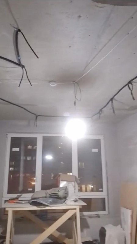 Комната в ходе ремонта с необработанным бетонным потолком и проложенными электропроводами, пластиковые окна и временный рабочий стол с инструментами.