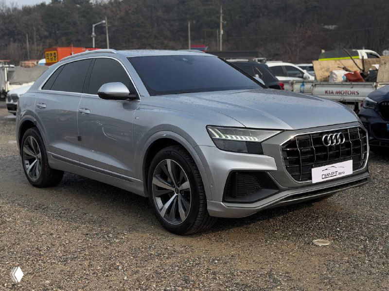 Audi Q8 50 TDI серого цвета, вид три четверти слева на парковке среди других машин, чистый кузов и стильные диски в фокусе.
