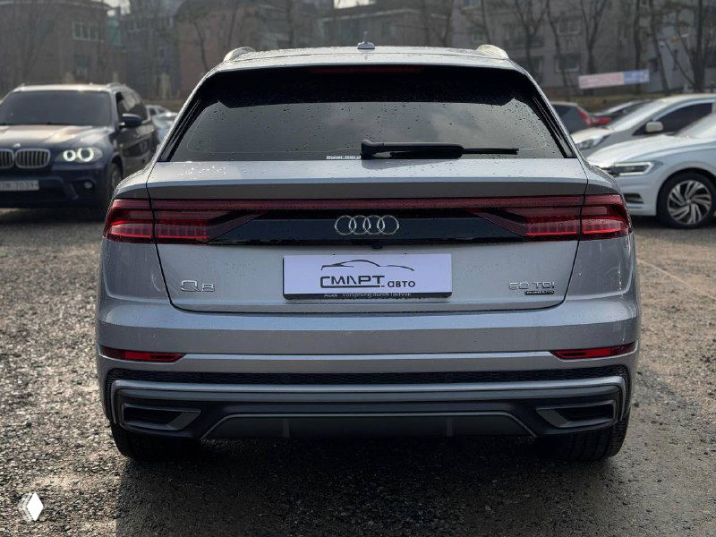 Вид на заднюю часть Audi Q8 с багажником и светодиодной полосой, серый кузов, парковка с рядом стоящими автомобилями в кадре.