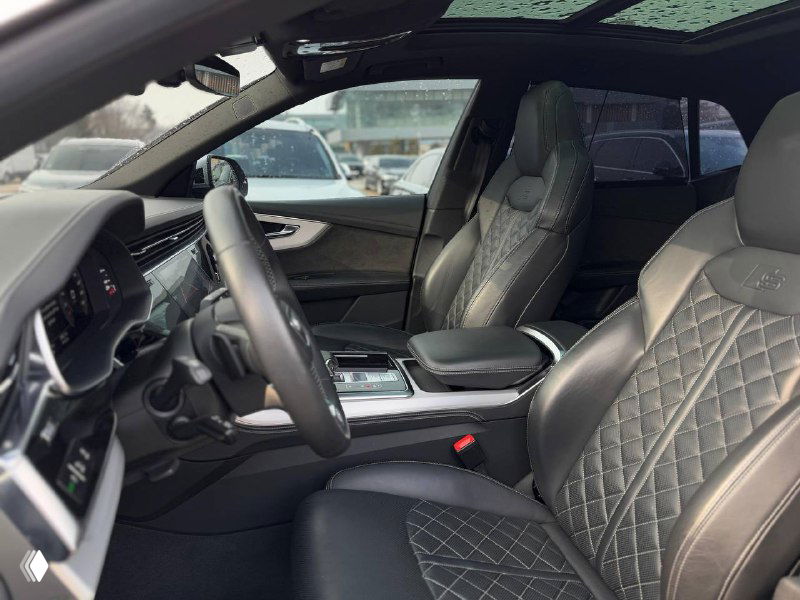 Вид на переднюю часть салона Audi Q8: водительское кресло, рулевое колесо, кожаная отделка и центральная консоль видны через раскрытую дверь.