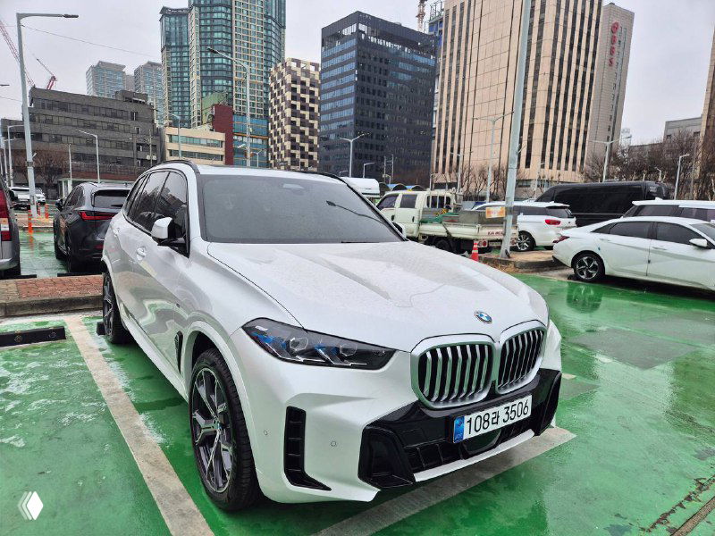 Белый BMW X5 30D xDrive M Sport, крупный план спереди и боковая часть на фоне высотных зданий, машина стоит на зелёной разметке парковки.