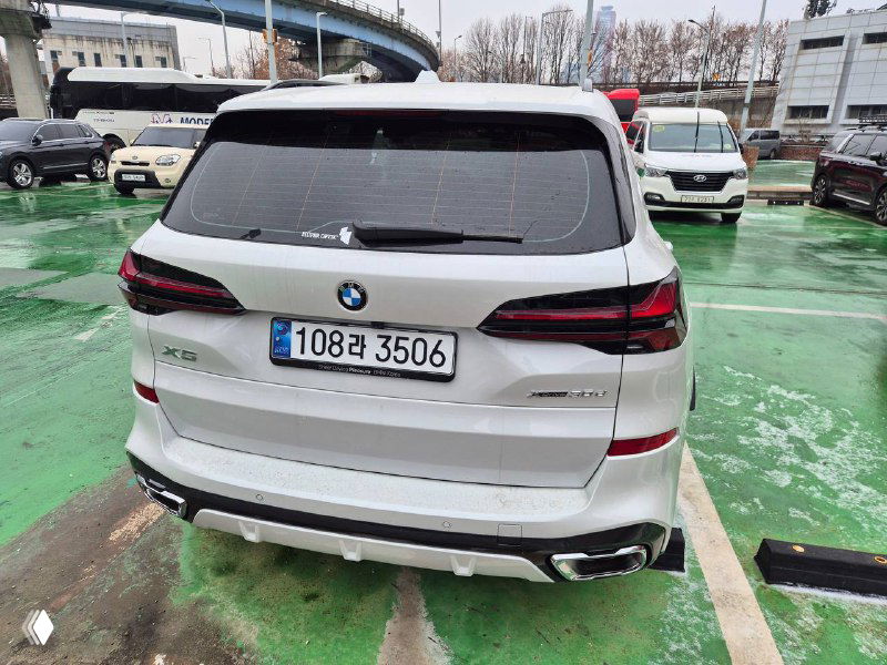 Вид сзади белого BMW X5 30D на парковке: задние фонари, бампер и номерная табличка, снег на земле и городские здания на заднем плане.