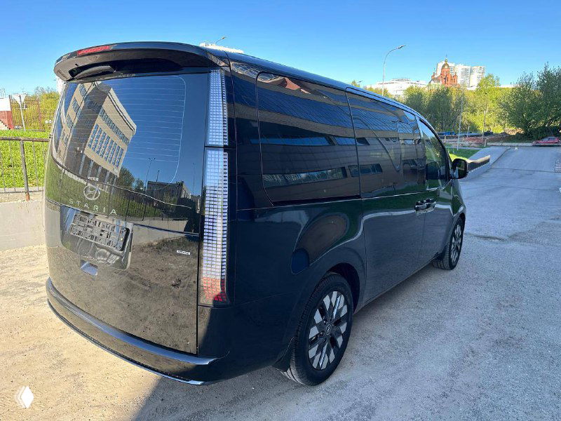 Задняя часть Hyundai Staria тёмного цвета, видно крупный фонарь и заднюю дверь, авто припарковано у многоэтажного здания.