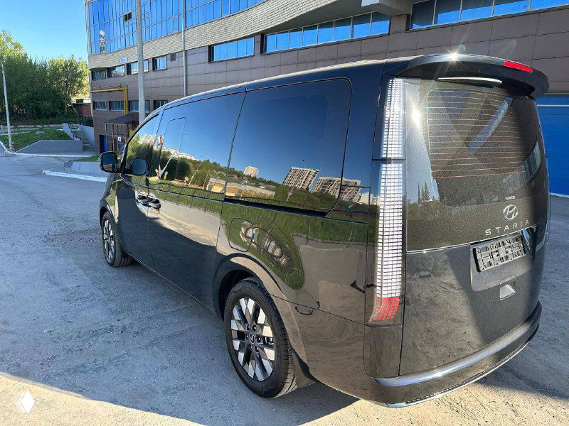Угловой задний ракурс Hyundai Staria, отражения зданий на лаковой поверхности кузова, видна форма задней части и боковины.