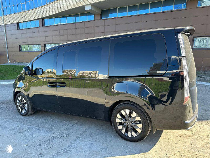 Боковой вид черного Hyundai Staria с пассажирской стороны, видны окна салона и контуры дверей на фоне городской архитектуры.