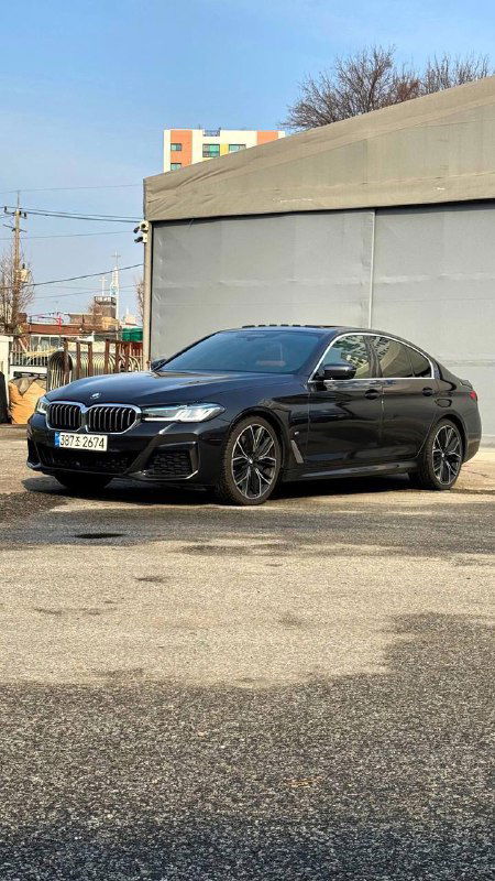 Боковой ракурс BMW 540i xDrive M Sport на асфальтовой площадке, видны диски, профиль кузова и чёрный цвет кузова на фоне здания.