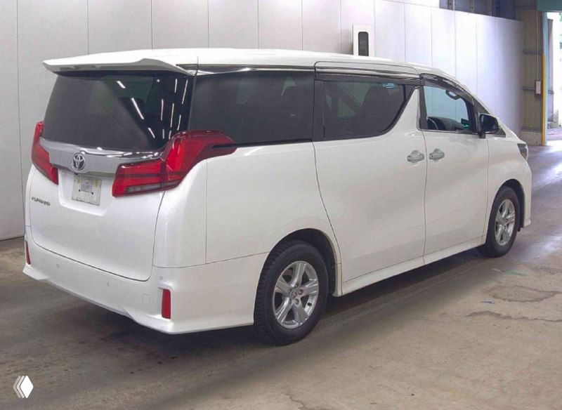 Toyota Alphard белого цвета — вид сзади и в профиль, задняя часть машины, фон ангарного помещения аукциона виден на фото, состояние кузова видно.