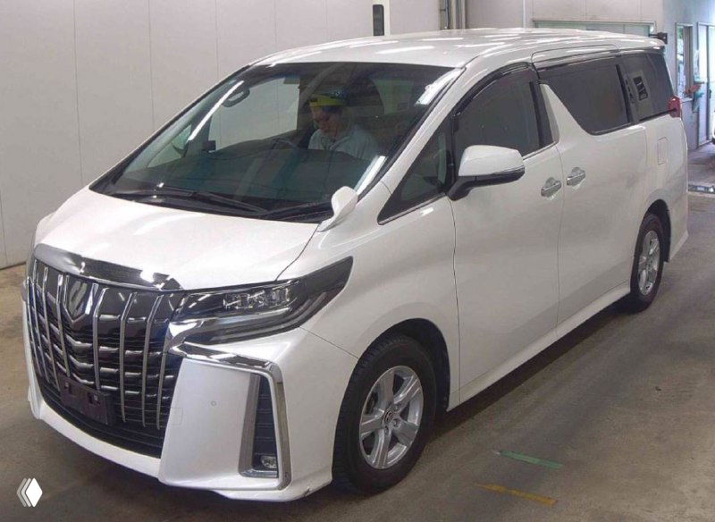 Диагональный фронтальный ракурс белого Toyota Alphard 2020 — капот, решётка, фары и боковая линия машины на освещённом фоне склада.