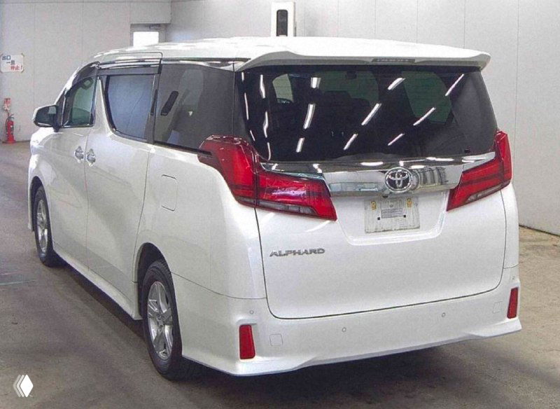 Вид сзади белого Toyota Alphard 2020 — открытая багажная дверь не видна, заметны задние фонари и эмблема, автомобиль стоит в помещении аукциона.