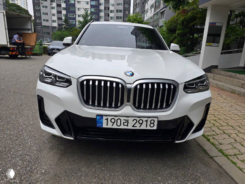 Крупный план передней части BMW X3 20i: двухсекционная решётка, фары и корейская номерная табличка на фоне двора жилого комплекса.