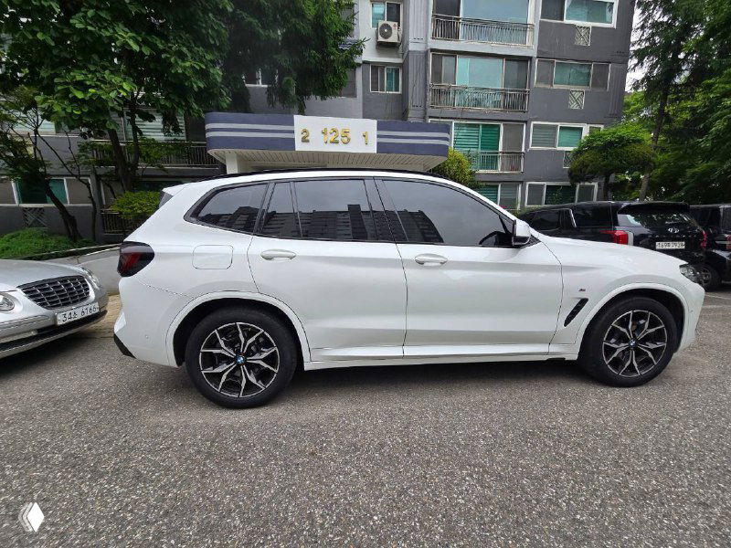 Профильная съёмка белого BMW X3 20i XDrive MSport в полный бок на фоне жилых зданий; видно боковые зеркала и линии кузова.