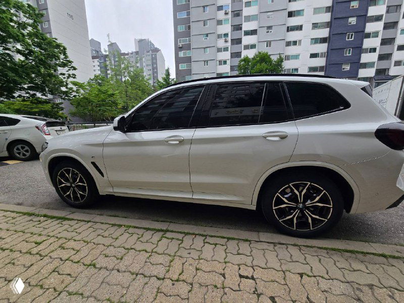 Боковой вид BMW X3 20i белого цвета, стоящий вдоль тротуара; видна длина кузова, колёсные арки и затемнённые стёкла.