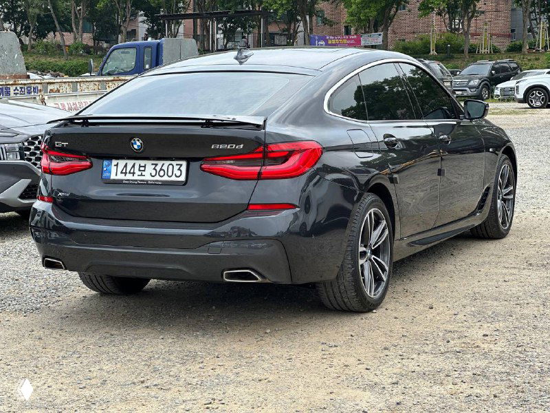 Задний вид BMW GT 620D XDrive: багажник, задние фонари и выхлопные насадки, автомобиль стоит на гравийной площадке, окружён грузовиками и машинами.