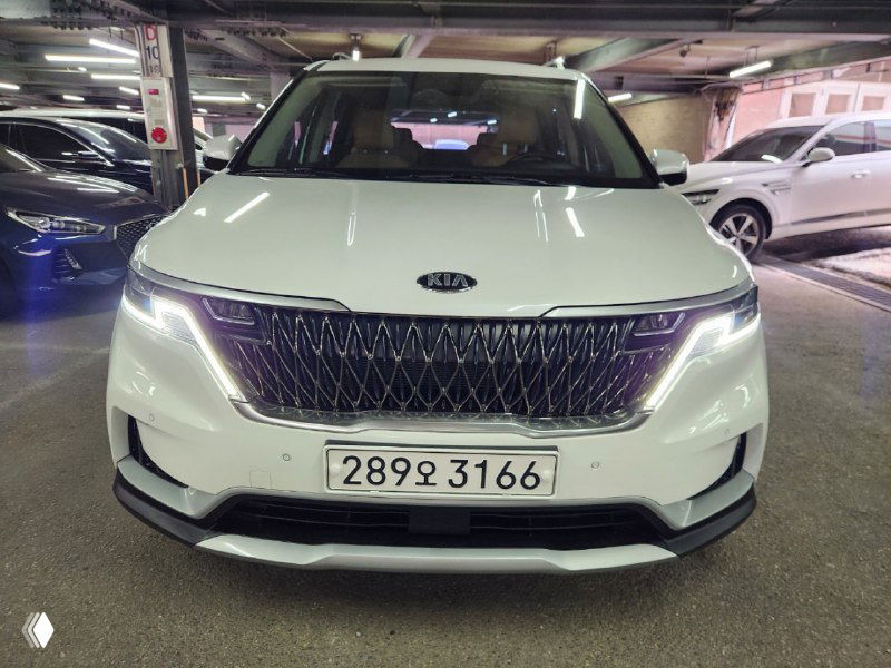Фронтальный вид белого KIA Carnival в паркинге, видна решётка радиатора, светодиодные фары и номерной знак корейской регистрации на автомобиле.