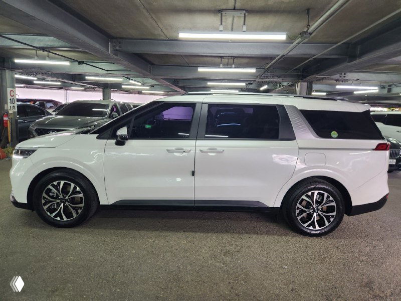 Профиль белого KIA Carnival в закрытом паркинге, заметна длина минивэна, боковые стекла и стиль дисков, автомобиль в стоянке дилера.