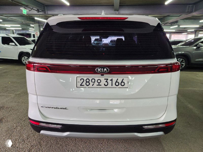 Задняя часть белого KIA Carnival с багажной дверью, эмблемой и задней светодиодной планкой; вид на номерной знак и шильдик модели.