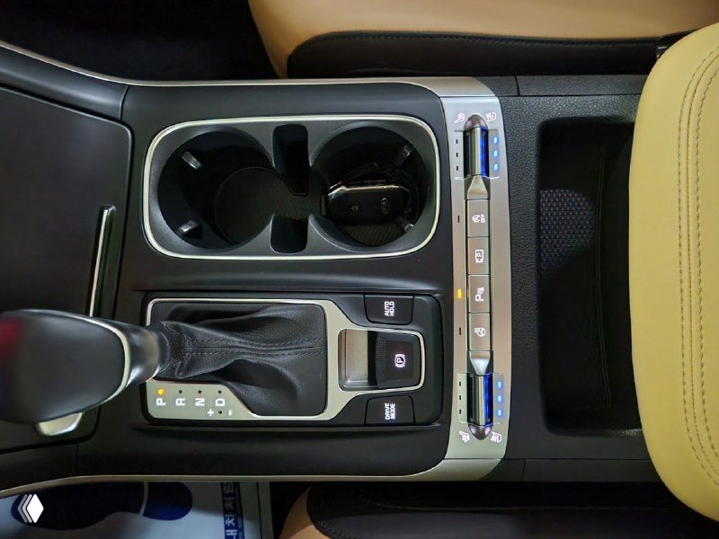 Крупный план центральной консоли KIA Carnival с шайбой переключения передач, подстаканниками и органами управления, видна отделка и элементы управления.