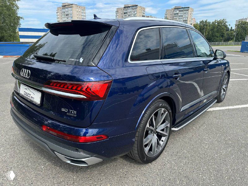 Задняя часть Audi Q7 тёмно‑синего цвета, видна оптика, бампер и эмблема Q7; фотография сделана под углом, городской пейзаж на заднем плане.