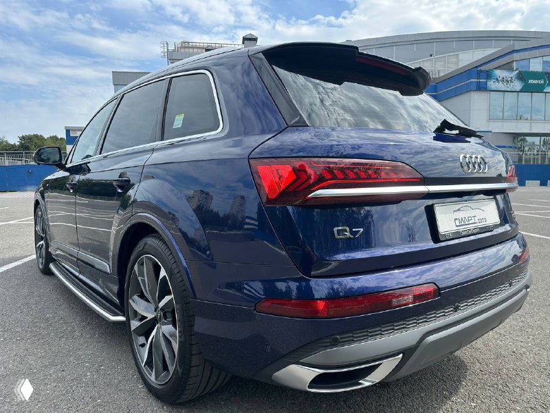 Крупный план задней стороны Audi Q7: задние фонари, эмблемы и выхлопные насадки, тёмно‑синий лак и отражения облачного неба.