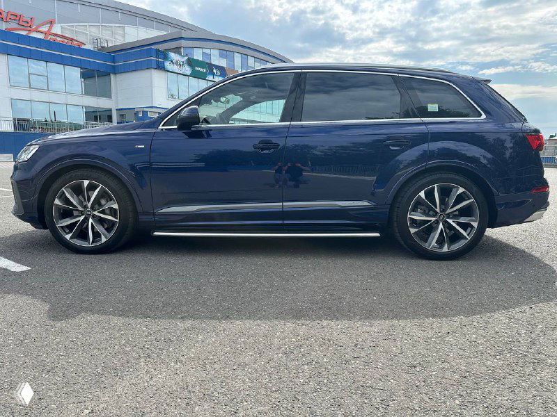 Сбоку уличный кадр Audi Q7, подчёркнуты диски и клиренс, заметны отражения зданий и голубое небо, автомобиль стоит на ровной асфальтовой площадке.