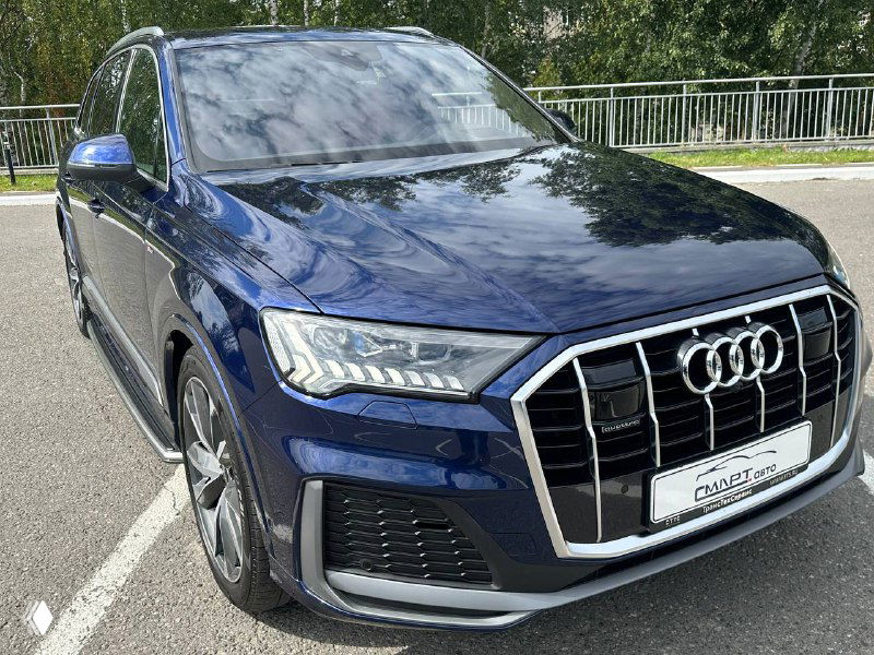Крупный кадр передней части Audi Q7, акцент на фальшрешётке, бампер и отражения неба на капоте; съемка подчёркивает премиальные элементы экстрерьера.