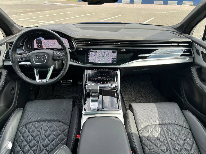Салон Audi Q7: передняя панель, мультимедиа‑экран и руль; видна отделка сидений, декоративные вставки и центральная консоль премиум‑уровня.