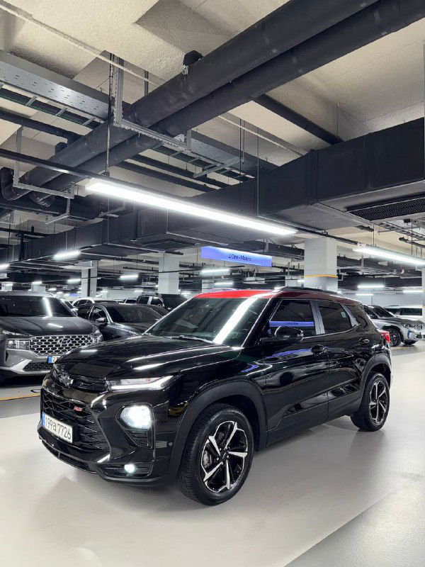 Общий трёхчетвертной ракурс Chevrolet Trailblazer чёрного цвета в паркинге: видно колёса, боковой профиль и отражения освещения на лаке.