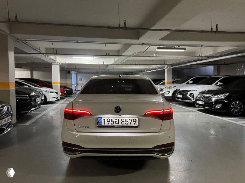 Вид сзади на белую Volkswagen Jetta в паркинге; заметна эмблема, габаритная светотехника и номерной знак автомобиля.