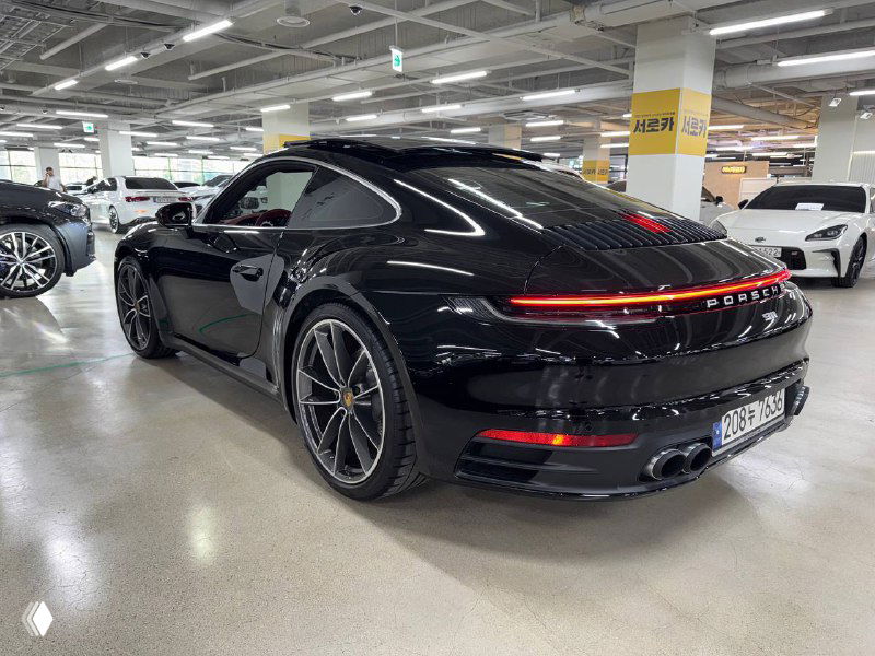 Тёмный Porsche 911 Carrera, общий ракурс сзади-вбок в крытой парковке; видно детали бампера и фон автосалона с колоннами.