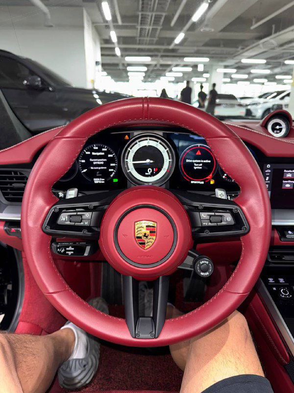 Рулевое колесо Porsche 911 с красной кожаной обивкой, логотипом Porsche в центре и цифровой приборной панелью позади руля.