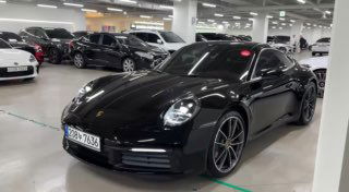 Чёрный Porsche 911 в крытом автосалоне на парковке: вид спереди‑слева, рядом другие автомобили и пространство шоурума вокруг, презентация лота.