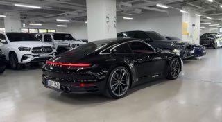 Задний вид чёрного Porsche 911 в автосалоне: LED‑полоса задних фонарей, форма кузова и диски на фоне других машин в шоуруме.