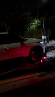 Белый автомобиль на автовозе ночью: частичный кадр кузова и фары при погрузке/транспортировке автомобилей по ночной дороге в городском окружении.