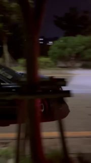 Чёрный седан частично виден на платформе автовоза ночью: задняя часть с огнями, городской фон и деревья вдоль дороги при транспортировке.