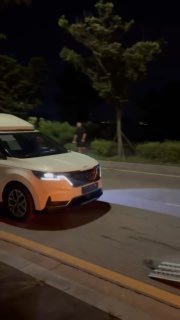 Белый минивэн (Kia Carnival) движется ночью по дороге рядом с автовозом, видна передняя часть, фары и уличное освещение при передаче партии.