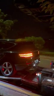 Тёмный внедорожник/кроссовер (Genesis GV80) на платформе автовоза ночью, видна задняя оптика и элементы погрузки рядом с обочиной дороги.