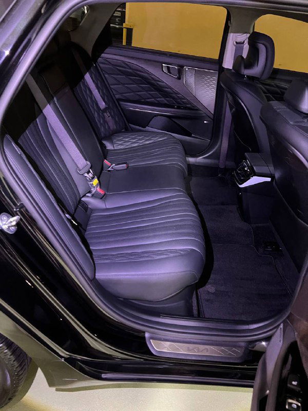 Вид на задние сиденья Kia K8 с открытой дверью: отделка кожей, ремни безопасности и чистый салон, фото сделано в паркинге